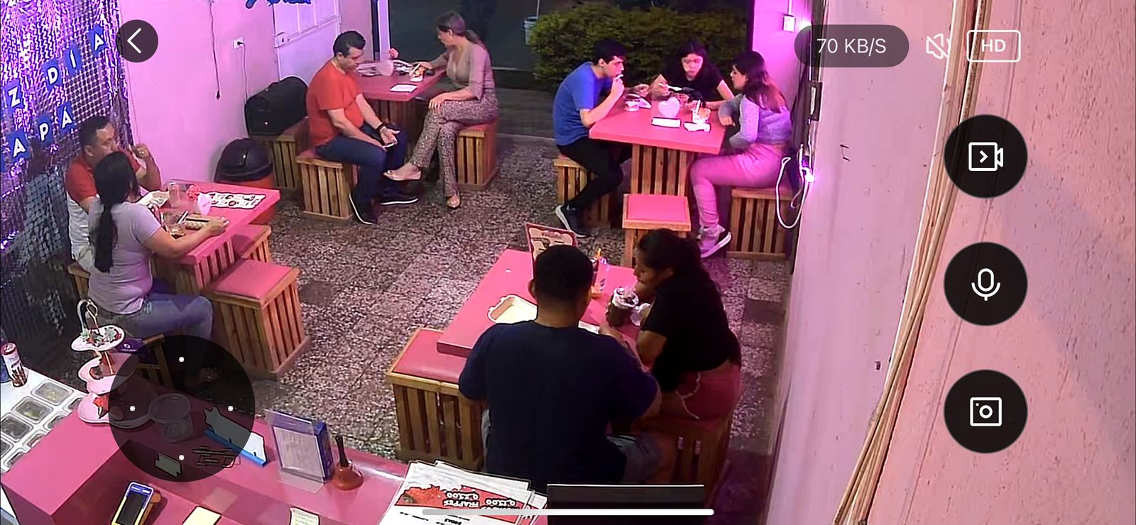 Una imagen de la perspectiva de una cámara de seguridad donde se miran varias personas sentadas alrededor de varias mesas, conviviendo unas con otras mientras disfrutan su comida